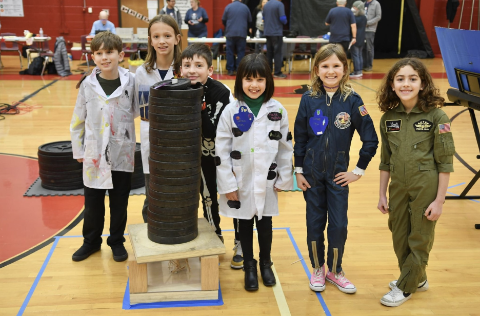 Image depicts six students in lab coats and flight suits smiling for the camera at the Odyssey of the Mind competition.